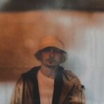 An artistic photo of a guy standing along a wall wearing a jacket, a white t-shirt, and a hat.